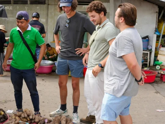 A traveler and guide buying fresh vegetables and herbs from a vendor at a traditional market for their cooking class.