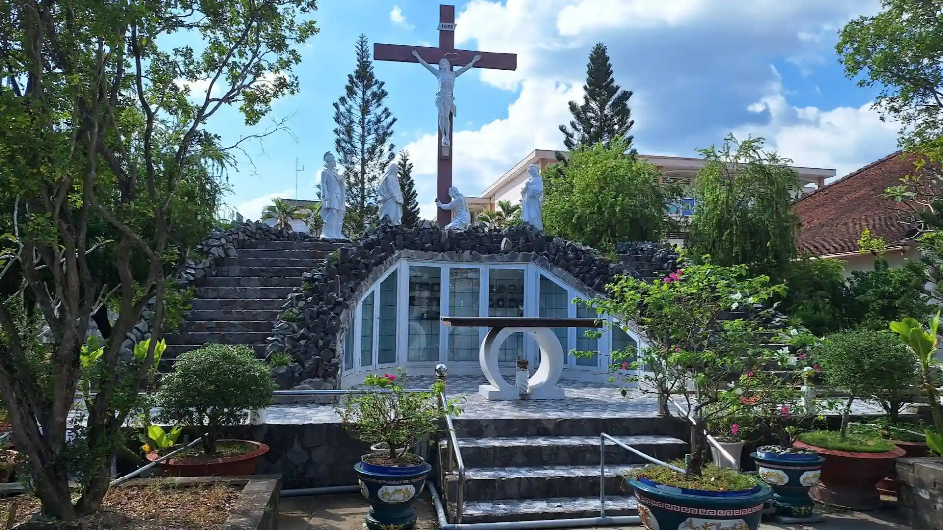 Outdoor Catholic shrine and crucifix at Cai Be Church, with potted plants and an open-air grotto