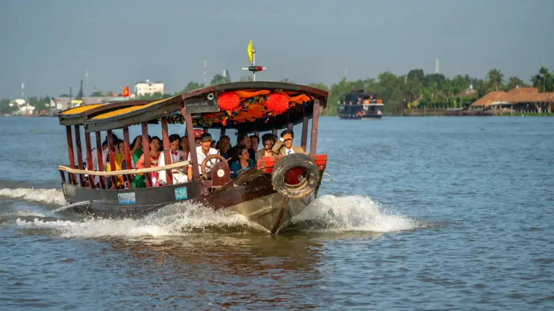 A wooden tourist boat filled with passengers cruising across the wide river toward Tan Phong Islet with Mekong Smile