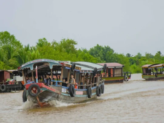 A traditional boat from the Me Kong Smile tour cruising along a wide river in Vinh Long